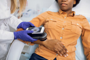 Preventive checkup, skin melanoma days concept. Close up of hands of female doctor dermatologist oncologist holding new generation dermatoscope, examining birthmarks and moles of afro lady.